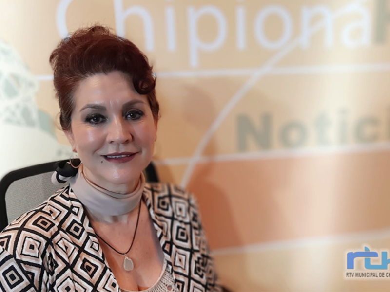 A una mujer con cabello rojo recogido, maquillaje suave y una blusa de cuadros. El fondo es borroso con letras en blanco y negro que parecen ser parte de un logotipo o marca. La imagen está firmada con "RTV.MUNICIPAL DE CHIPIONA" en la esquina inferior derecha.