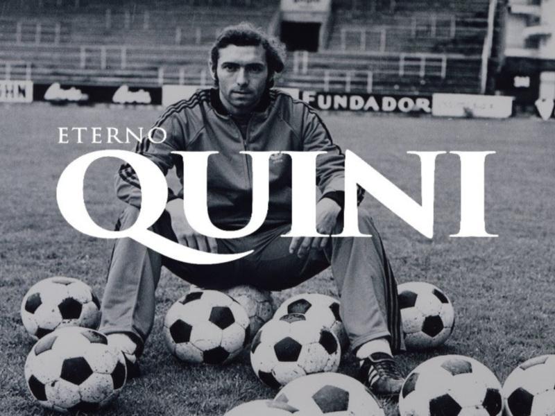 Un hombre sentado en el campo de fútbol, rodeado de balones de fútbol. La imagen es en blanco y negro con letras blancas que dicen "ETERNOS QUININI".