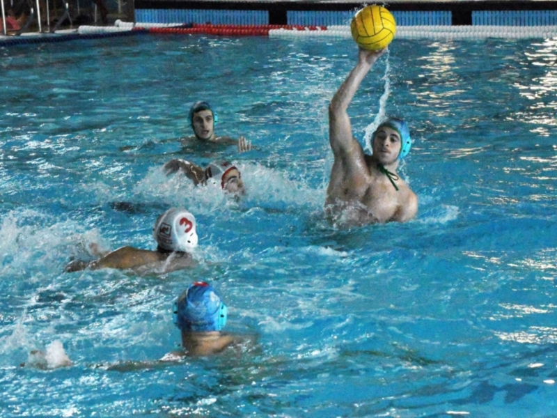 Jugadores de waterpolo en acción en una piscina de agua azul, con un jugador del equipo azul preparándose para lanzar la pelota amarilla en el aire.