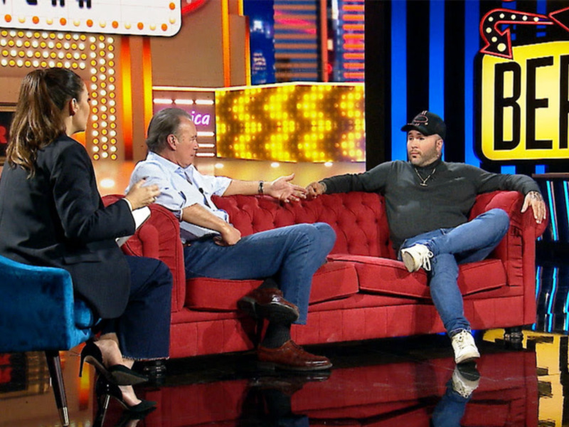 Una imagen de una conversación en un programa de televisión, con tres personas sentadas en sillones rojos. La escena incluye un fondo con luces y una pantalla con texto.