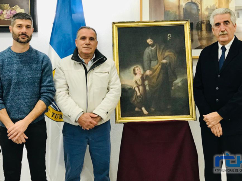 Dos personas posan junto a una pintura religiosa en un entorno formal, con una bandera y una obra de arte en el fondo.