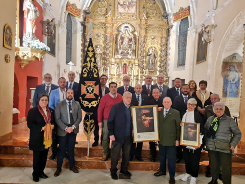 Una imagen de una iglesia con un altar y estatuas. En el centro, hay una persona sosteniendo un cartel dorado con un retrato de un hombre. Alrededor, varios adultos están posando para la foto, algunos con chalecos y otros con abrigos. La imagen parece ser una celebración o reconocimiento en la iglesia.