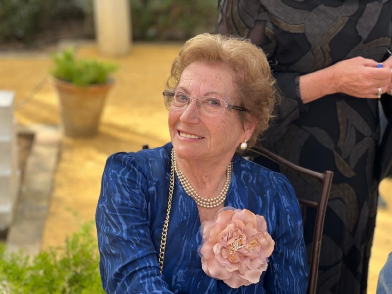 Retrato de una mujer mayor sonriente, vestida con un traje azul y una flor en el pecho, sentada en un patio soleado.
