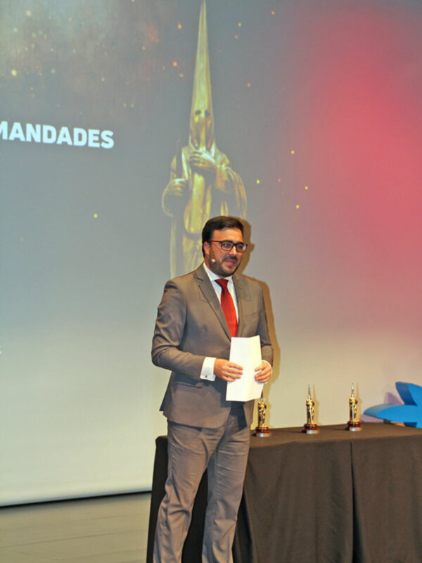 Hombre en traje gris presentando ante un escenario con un trofeo en la mesa.