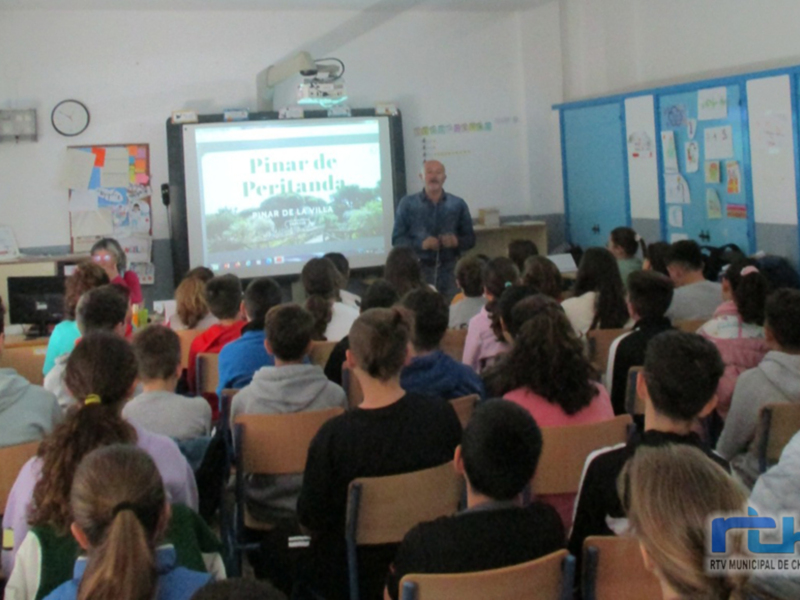 Estudiantes en una aula escuchando a un profesor frente a un proyector con la imagen de "Pinar de Peritanda". La sala está decorada con carteles y papeles colgados en las paredes.