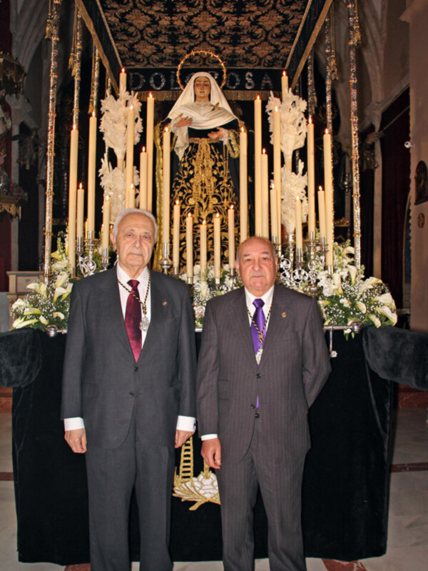 Dos hombres en trajes posan frente a una estatua de la Virgen María con velas y flores en un altar.