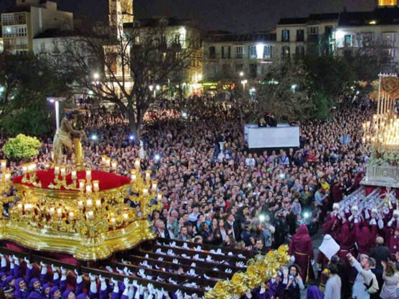 Cientos de personas participan en una procesión nocturna, con dos altares iluminados y una multitud de participantes vestidos en trajes tradicionales. La escena se desarrolla en un entorno urbano con edificios y árboles alrededor.