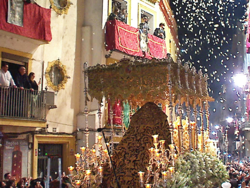 Festividad religiosa en la calle, con una procesión de imágenes en el centro y personas observando desde los balcones.