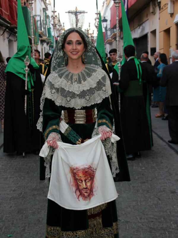 Una mujer en traje tradicional con un vestido verde y negro, llevando una falda larga negra con detalles dorados. Su rostro está cubierto por un velo verde, y sostiene una bandera blanca con un rostro pintado. La imagen muestra a otros individuos en trajes similares, caminando por una calle con edificios coloridos a ambos lados.