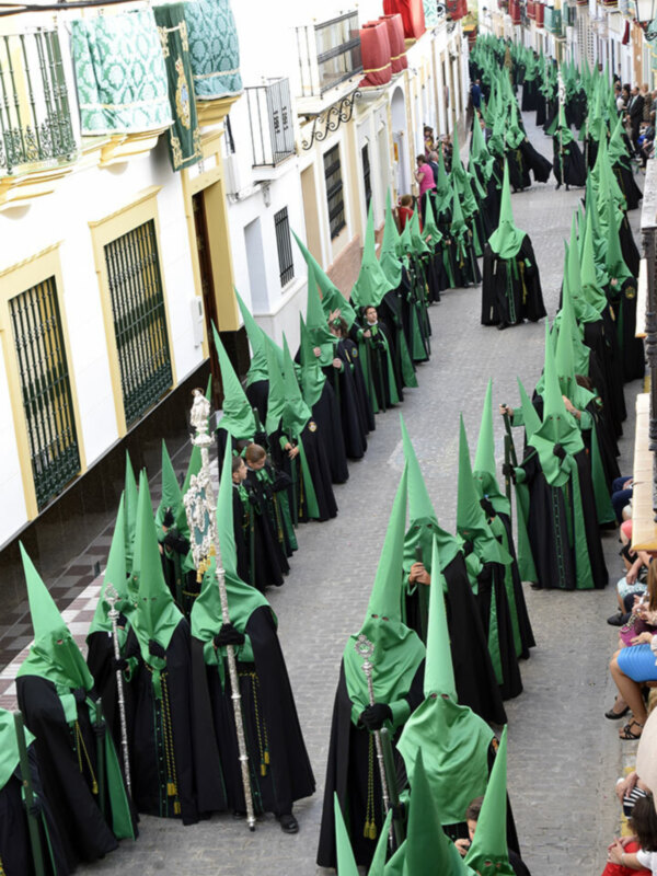 Mujeres vestidas con trajes tradicionales de la Semana Santa, portando capuchas verdes y mantos negros. Celebración religiosa en una calle de la ciudad, con gente observando desde los balcones.