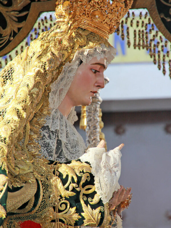 Una estatua de oro y plata con detalles elaborados, posiblemente una Virgen María, vestida con un manto de seda y oro. La estatua está en un entorno arquitectónico con adornos dorados y cristales.
