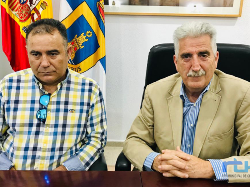 Dos hombres sentados frente a una mesa con banderas españolas y regionales en el fondo.
