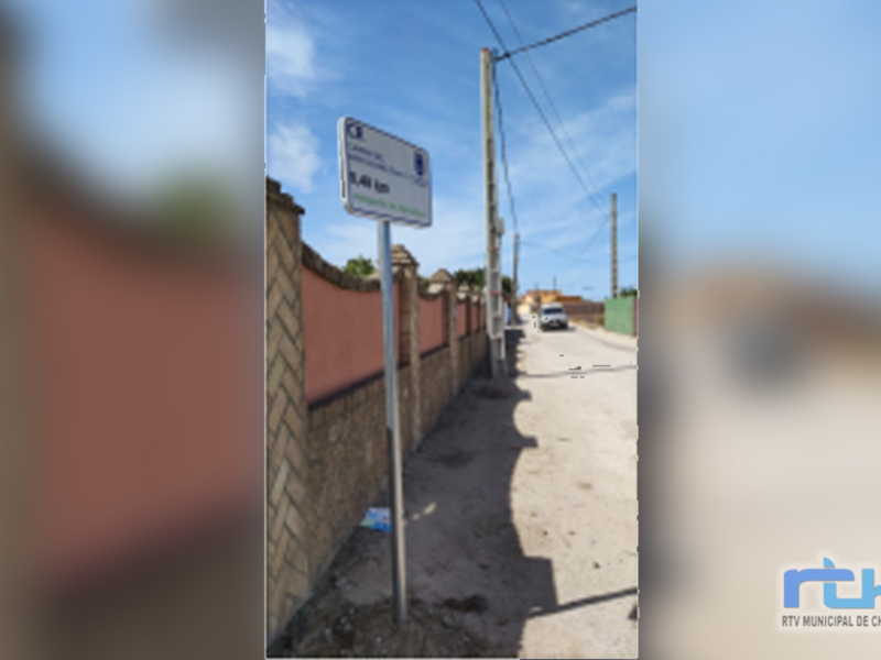 Vista de una calle con una pared de ladrillos y un poste de electricidad. El cielo es azul claro y hay un vehículo en la distancia. La imagen parece estar tomada en un área residencial o urbana.