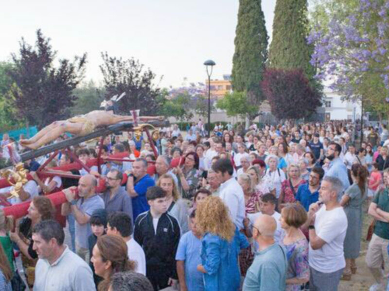 Una multitud de personas congregadas en un evento, con un grupo de personas en el centro, seguramente en un acto religioso o festivo, rodeadas por árboles y una estructura roja en el fondo.