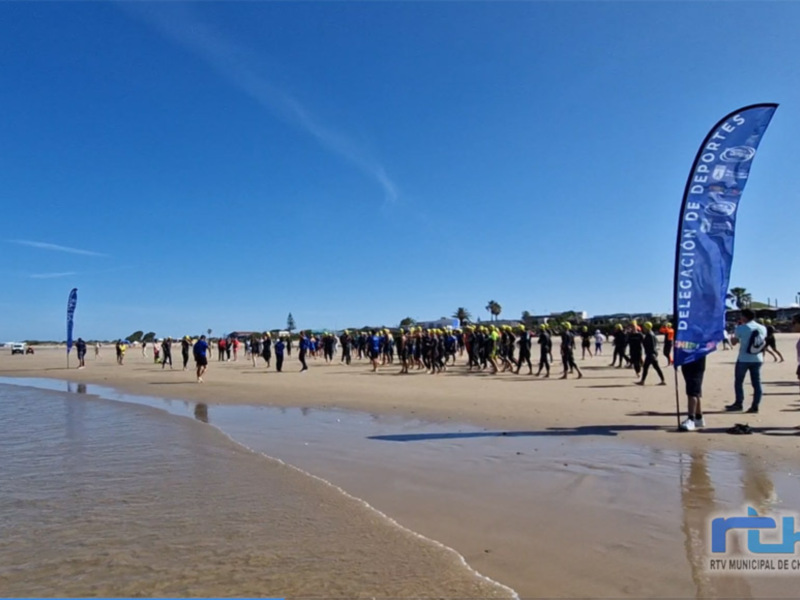 Desfile de personas en una playa con banderas y un cielo azul claro. La delegación de deportes está presente con una bandera azul en la playa.