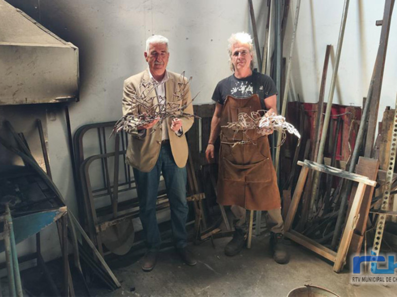 Dos hombres posan con ramas de árbol en una oficina o taller, rodeados de herramientas y equipos de trabajo.