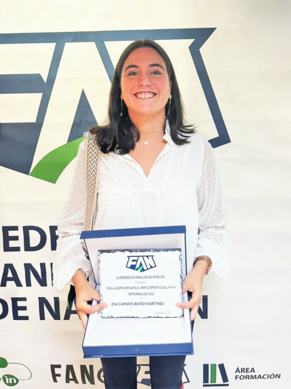 Una mujer sonríe mientras sostiene un diploma de la Federación Andaluza de Natación, celebrando su reconocimiento deportivo.