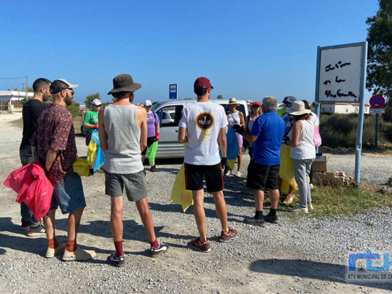Un grupo de personas con mochilas y ropa casual se preparan para un evento al aire libre. En el fondo, hay una caravana y un cartel con instrucciones para los visitantes. La escena está iluminada por el sol, y la naturaleza circundante es visible.