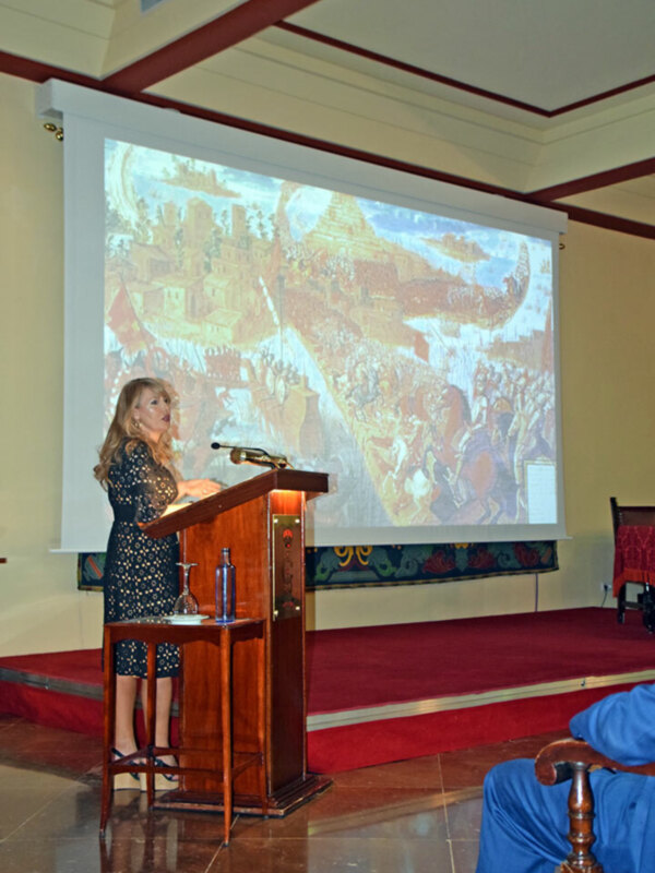 Una mujer en un vestido azul se encuentra detrás de una mesa de conferencia, con un proyector en la pared y un mapa del mundo en el fondo.