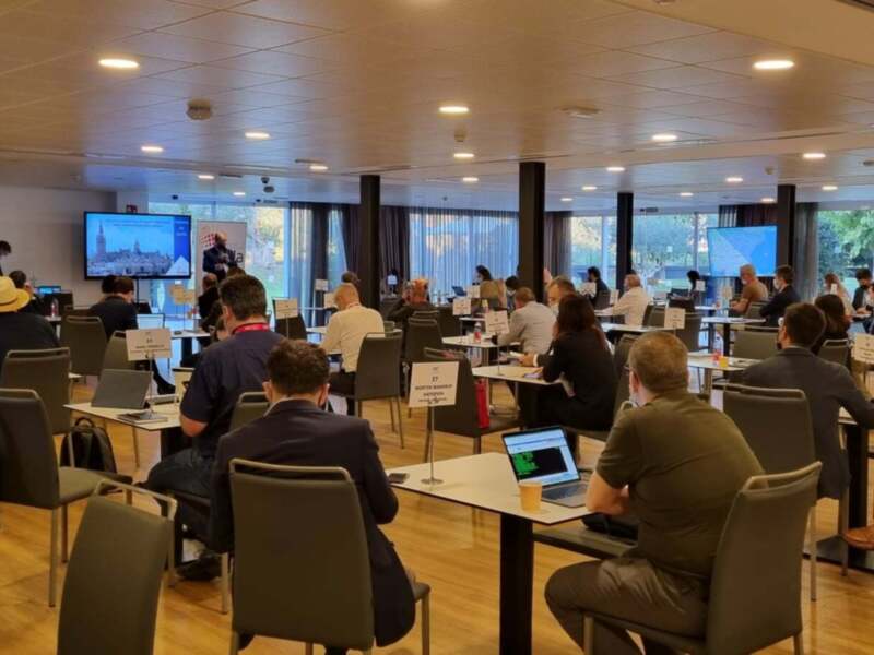 En una sala de conferencias moderna, varios participantes se encuentran sentados en sillas frente a computadoras. La sala está iluminada con luces de techo y cuenta con grandes ventanas que permiten la entrada de luz natural. La decoración es minimalista, destacando el uso de madera y la estructura metálica del techo.