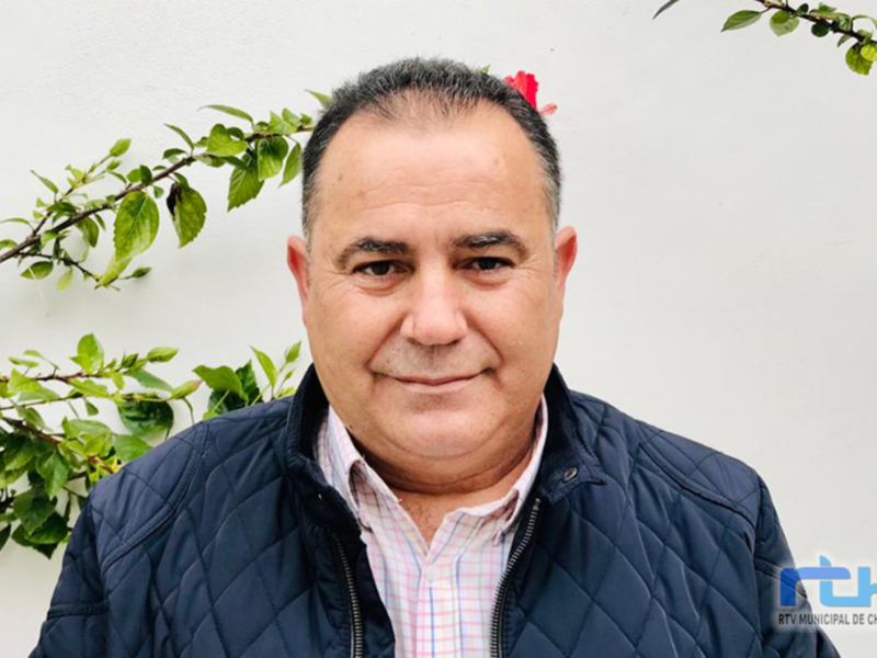 Retrato de un hombre con una chaqueta azul y una camisa a cuadros, con un ramo de flores en el cabello, frente a un fondo blanco con hojas verdes en la parte superior.
