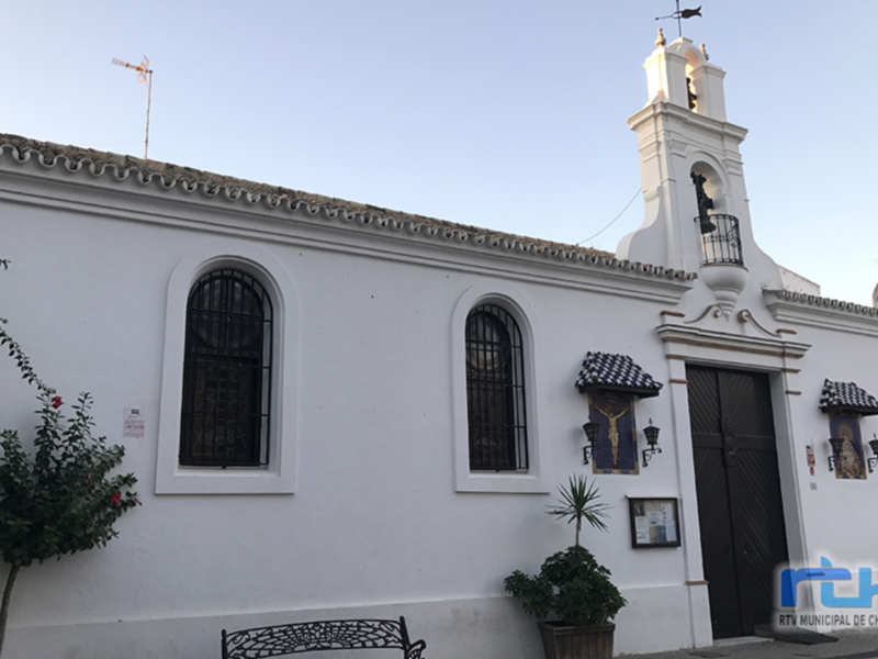 Una imagen de una iglesia blanca con detalles en blanco y negro. La torre tiene un campanario, una bandera azul y negra en la fachada. Hay ventanas con persianas, una puerta principal y un banco de metal en la entrada. La arquitectura es clásica, con detalles geométricos y una chimenea en la parte superior. La imagen parece ser de un pueblo o ciudad histórica, posiblemente en España.