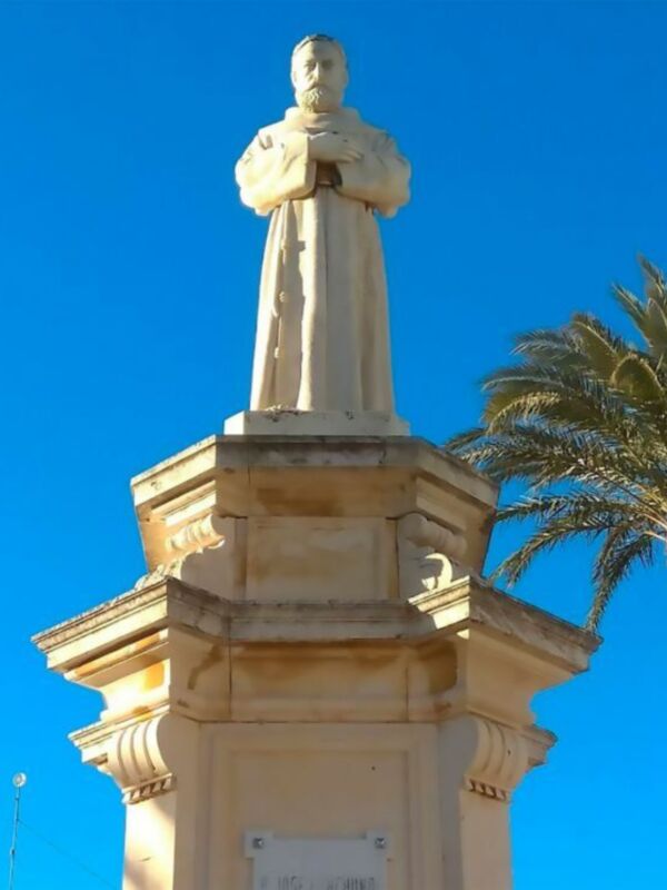 Estatua de un hombre con capa blanca en una base de piedra, contra un cielo azul claro. A la derecha, se ve una palmera alta y verde. En el pedestal hay una placa con texto.