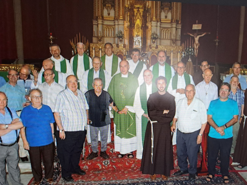 Una imagen de una iglesia con un grupo de personas posando juntos. La gente está vestida formalmente, muchos llevan camisas y pantalones a juego. Al fondo se ve una gran imagen de la Virgen María en un altar, rodeada por otros santos y figuras religiosas. La imagen está tomada en una iglesia con un techo alto y columnas doradas.