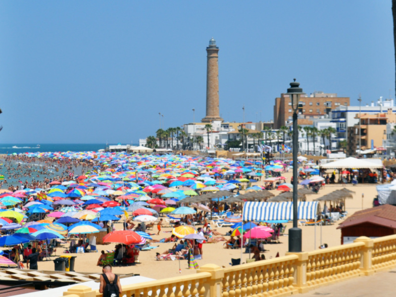 Playa de la Barrosa en Cádiz, España. Un día soleado con numerosos visitantes disfrutando del clima y la belleza de la playa. En el fondo, se puede ver el faro de la Barrosa, una estructura icónica de la ciudad.