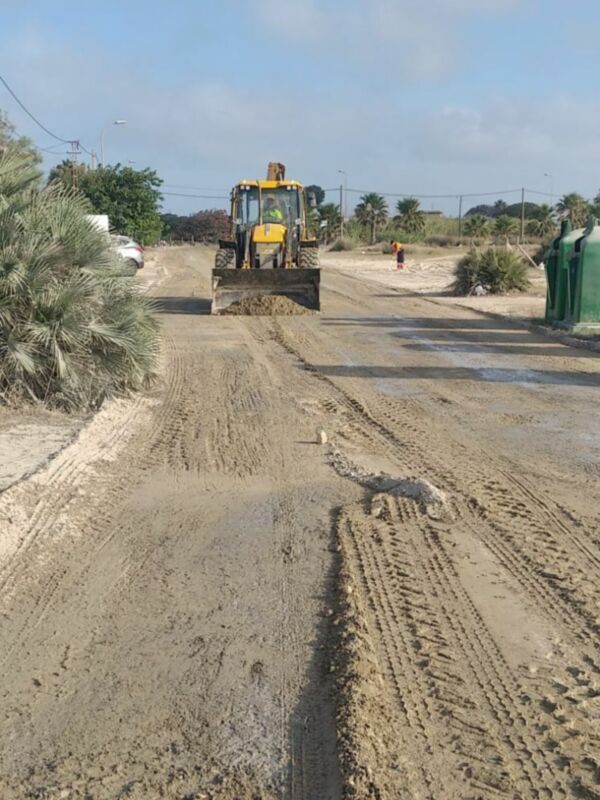 Una excavadora amarilla trabajando en un camino de tierra, con árboles y una carretera al fondo.