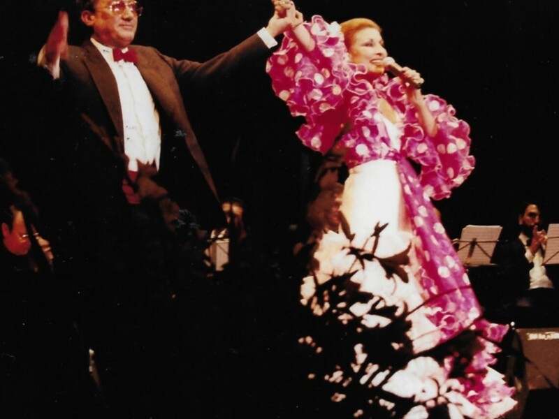 Flamenco dancer in vibrant attire performing on stage with a conductor in the background.