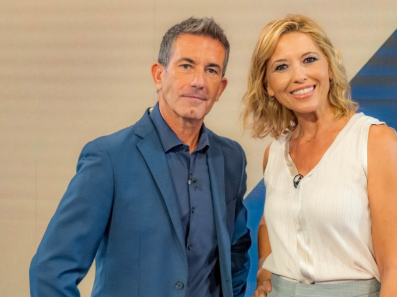 Dos personas posan juntas en un entorno profesional, uno con traje azul y la otra con vestido blanco, frente a una pantalla de fondo.