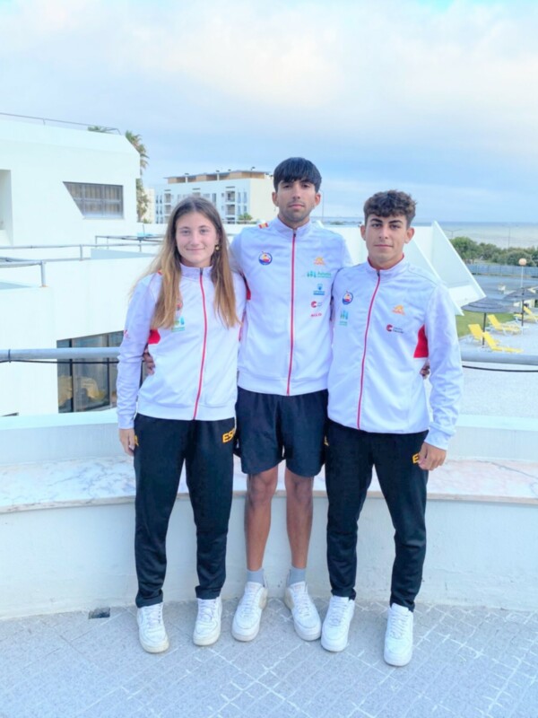 Tres jóvenes atletas posan para una foto en una terraza con vistas al mar, vestidos con uniformes deportivos blancos y negros.
