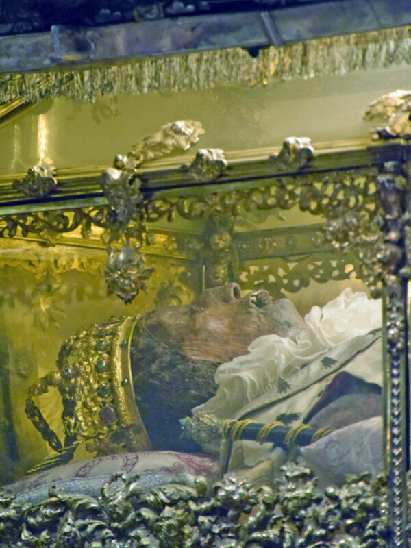 Una escultura de un personaje histórico, posiblemente un rey o monarca, vestido con una corona y sosteniendo un libro. La escultura está rodeada por detalles dorados y ornamentados, sugiriendo un entorno de nobleza o religión.