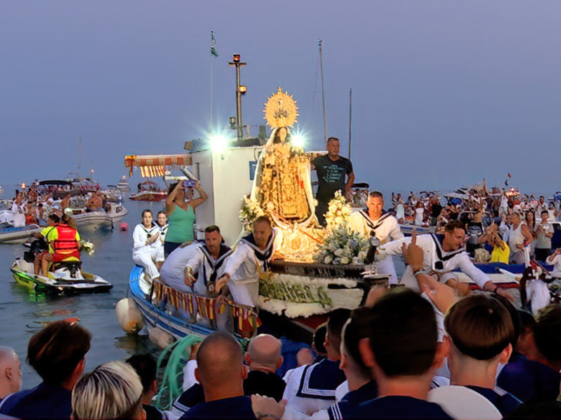 Una procesión religiosa en un puerto con una imagen de la Virgen en el centro, rodeada por personas vestidas con trajes tradicionales y banderas ondeando.