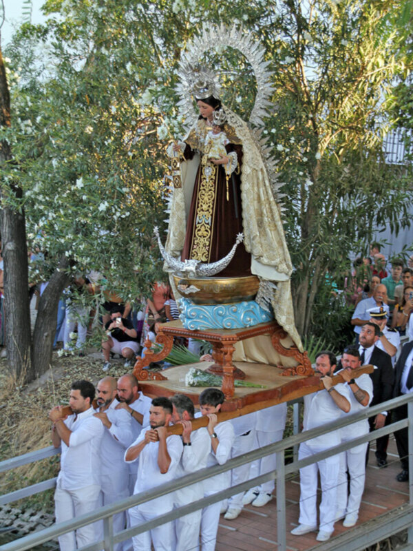 Procesión de la Virgen con portadores en traje blanco, en un entorno natural con árboles y flores.