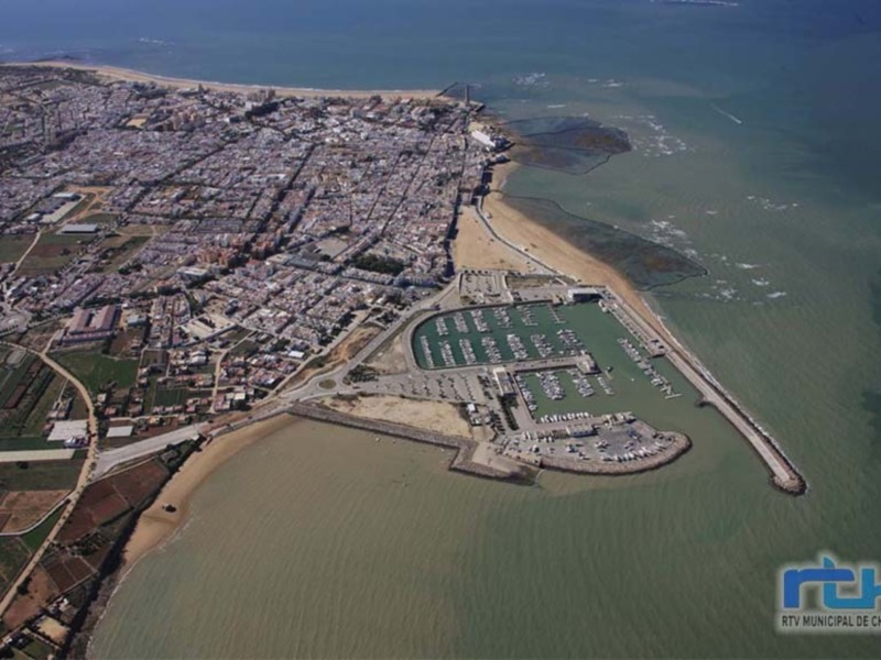Imagen aérea de Chipiona, una ciudad costera en España. Se observa el puerto deportivo y la playa a lo largo de la costa. La imagen muestra una mezcla de urbanización y naturaleza, con edificios, jardines y el mar.