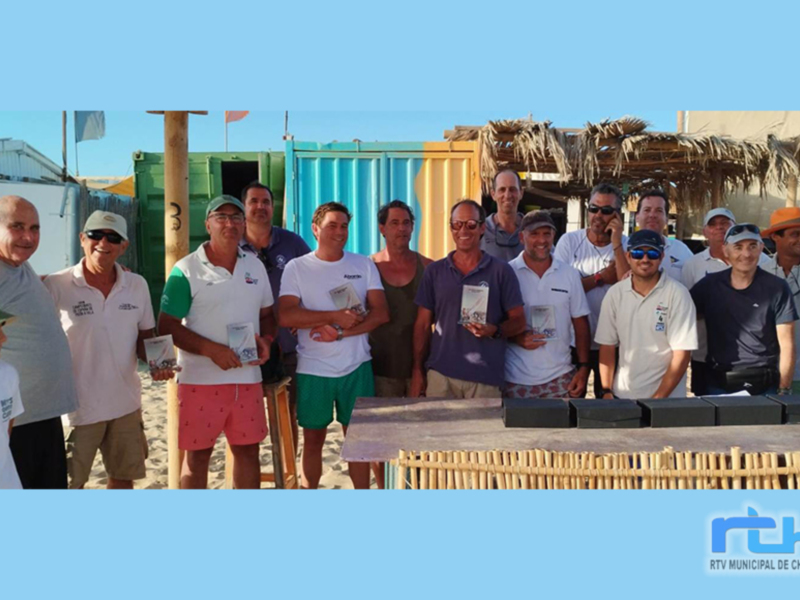 Equipo de personas posando para una foto en la playa, con un fondo de contenedores y una estructura de palma. El equipo parece estar celebrando un evento deportivo o social.