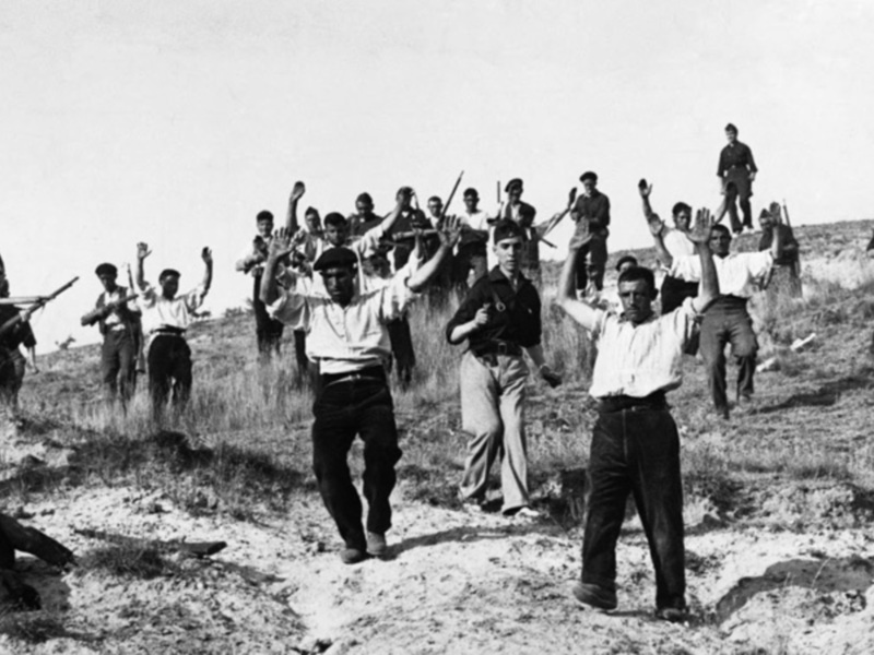 A un grupo de personas en una escena histórica, posiblemente durante la Guerra Civil Española. Las personas están levantando las manos, lo que podría indicar un gesto de rendición o protesta. La escena se realiza en una zona abierta con un cielo despejado, lo que sugiere que la acción está ocurriendo al aire libre. La imagen es en blanco y negro, lo que aporta un tono histórico y dramático al momento captado.