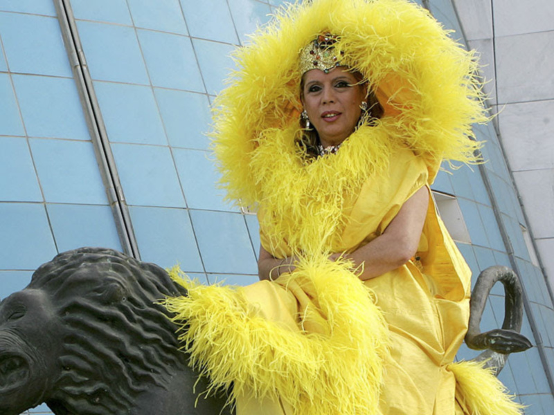 Una mujer vestida de amarillo con plumas y un sombrero grande, posando en una estatua de león negro.