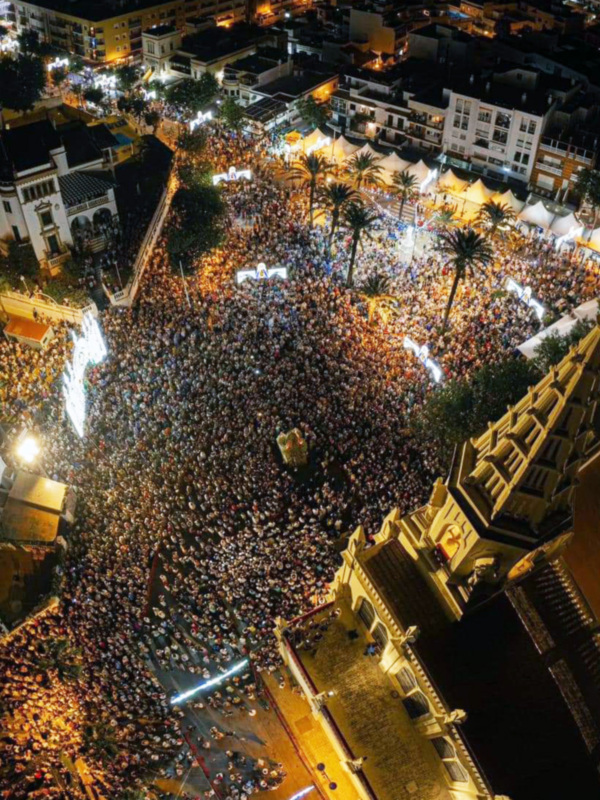 Festividad nocturna en la ciudad, con multitud de personas reunidas en la calle. Palacios y edificios antiguos rodean la celebración. La iluminación festiva ilumina el ambiente.