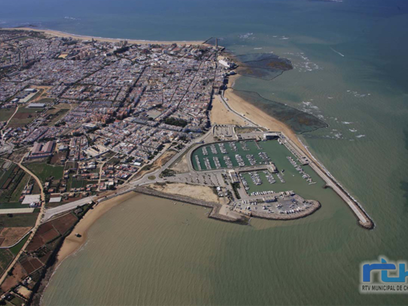 Imagen aérea de Chipiona, una ciudad costera en España. La imagen muestra la costa con playas y edificios urbanos, así como un puerto deportivo cercano a la ciudad.