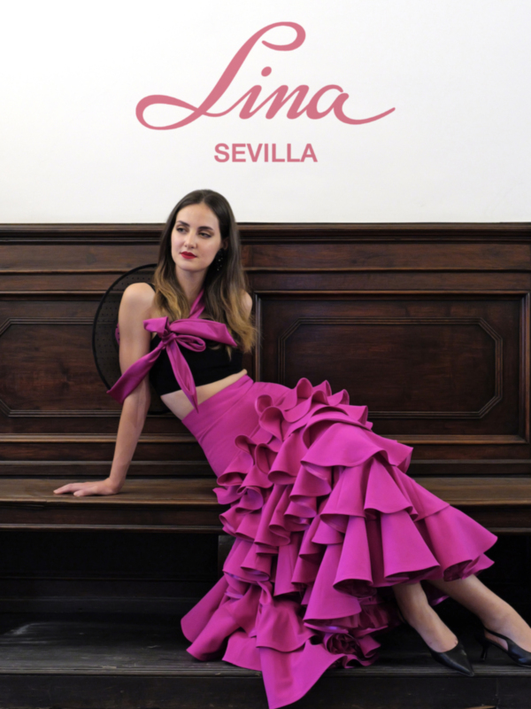 Model wearing a vibrant pink ruffled dress by Lina Sevilla, posing elegantly on a wooden bench.