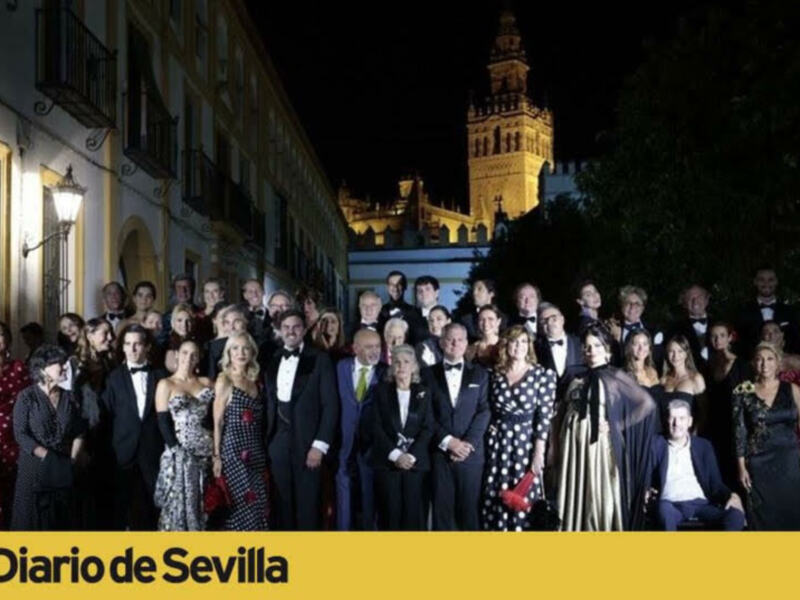 Grande grupo de personas en trajes formales posando para una foto en la noche, con el fondo de la torre de la Giralda de Sevilla iluminada.
