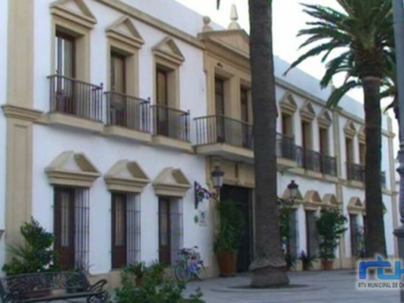 Fachada de un edificio colonial en una plaza con palmeras y bancos.