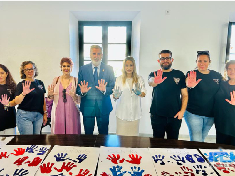 Una imagen de una reunión en un espacio moderno con personas de pie detrás de una mesa. La mesa tiene varios sellos de manos en diferentes colores, y las personas están mostrando sus manos. La imagen transmite un mensaje de inclusión y diversidad.