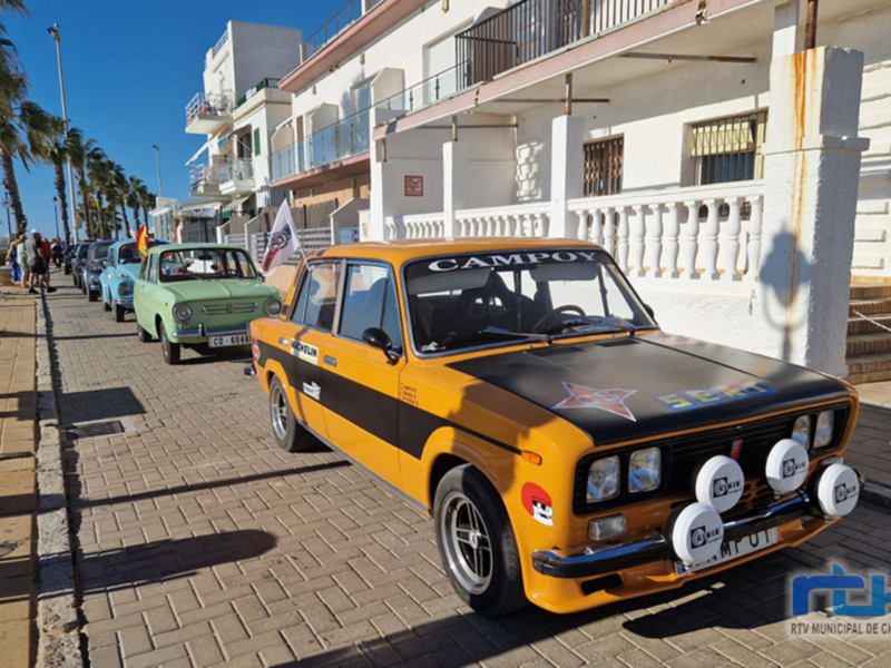 Vista de una calle con coches antiguos estacionados frente a edificios blancos. El coche amarillo con el texto "CAMPO" en la parte superior es el más destacado. La imagen muestra un día soleado con sombras de los coches y edificios en el suelo.
