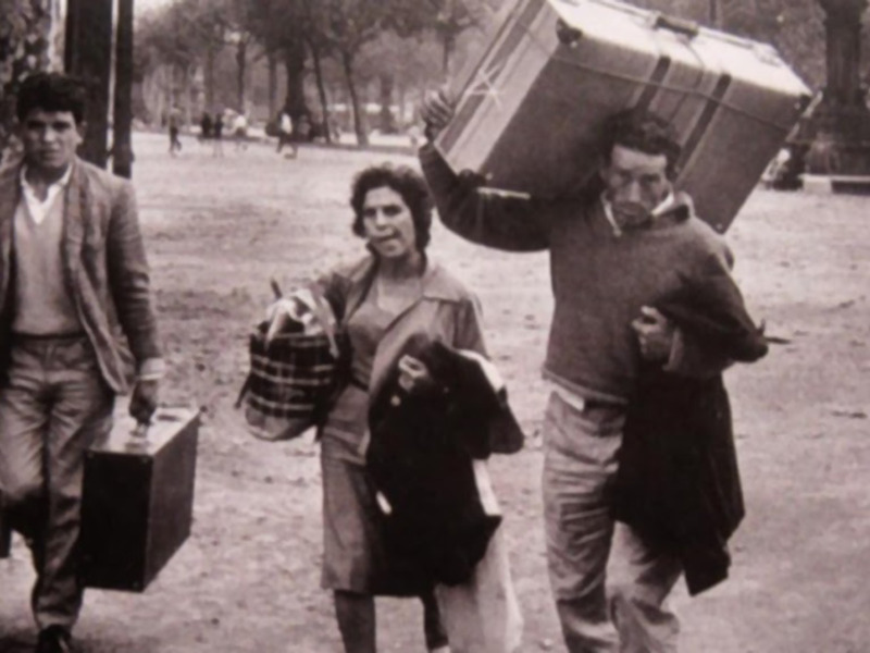 Una familia de tres personas camina por un parque con maletas y bolsos. Parece ser una escena de desplazamiento o emigración, posiblemente en la década del 70.