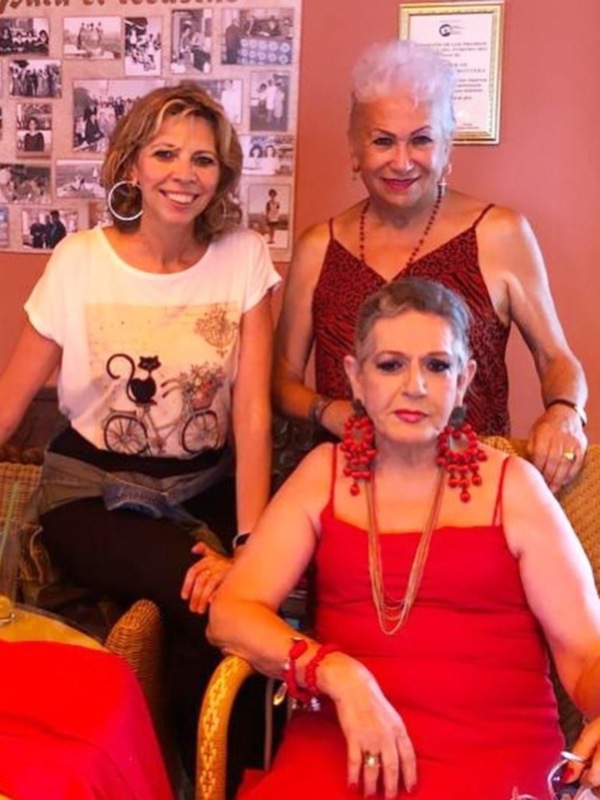 Tres personas sentadas en una mesa, con una mujer en el centro vestida de rojo y dos mujeres a su lado, una de ellas con una camiseta con un dibujo de una bicicleta y gato.