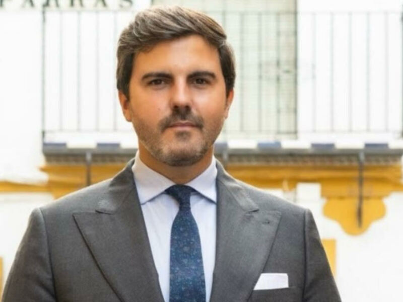 Elegante hombre en traje y corbata, posando frente a una estructura de metal con detalles amarillos.